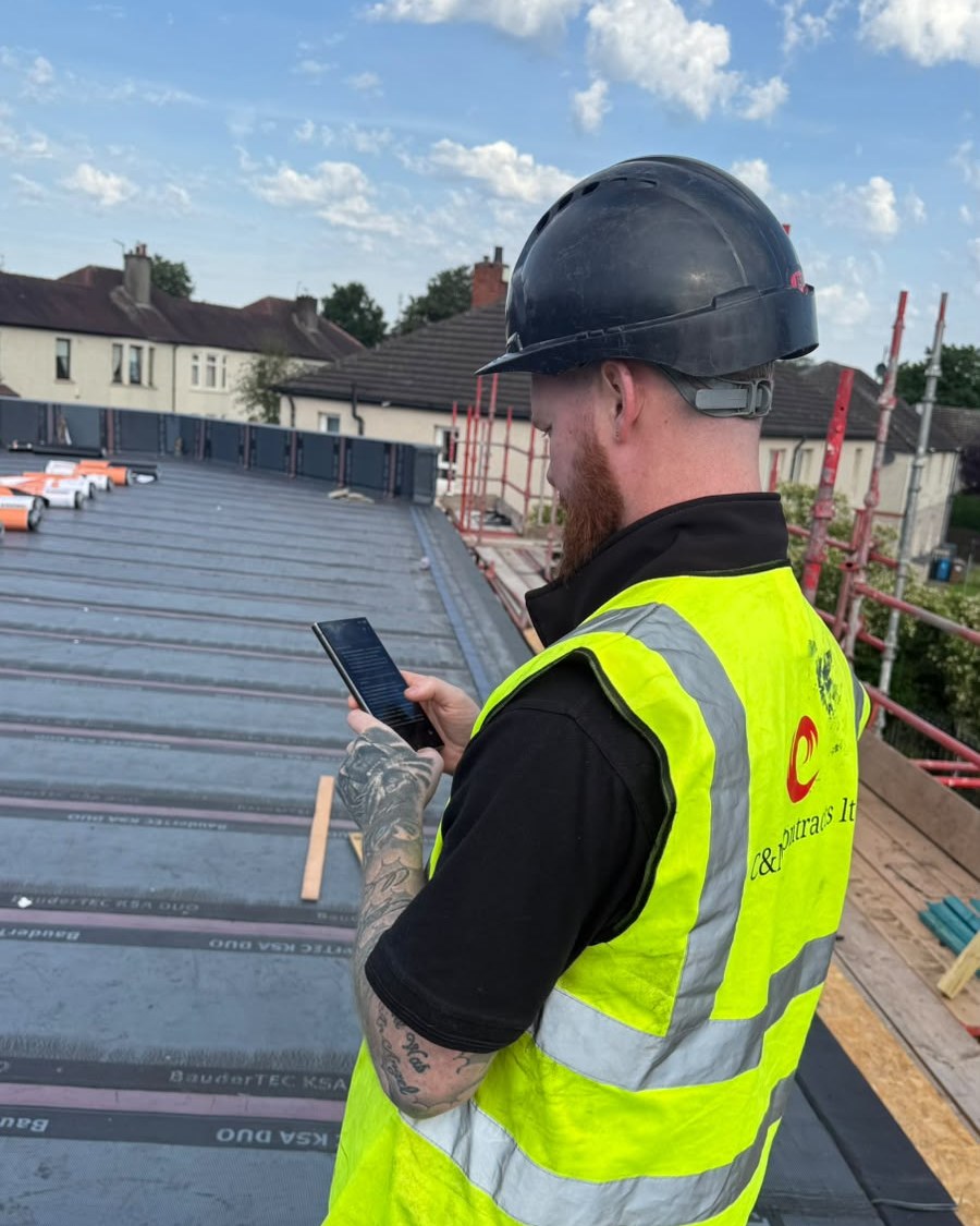 UK roofing contractor in hi-vis and hard hat using the RunwiseOS app on his phone, on a flat roof mid-installation with scaffolding and rooftops behind